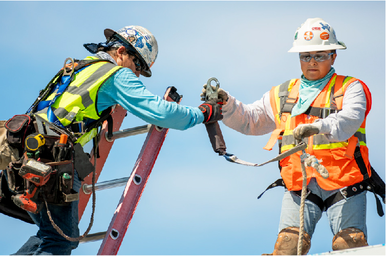 Two construction workers wearing safety gear and helmets, working together on a ladder and rooftop, securing a safety harness
